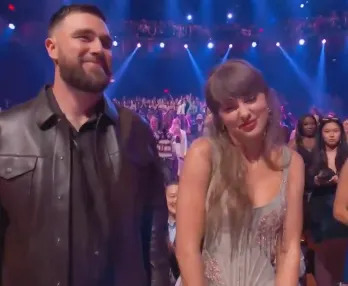 Two people stand together at an event with a crowd behind them. One is in a leather jacket, the other in an elegant gown