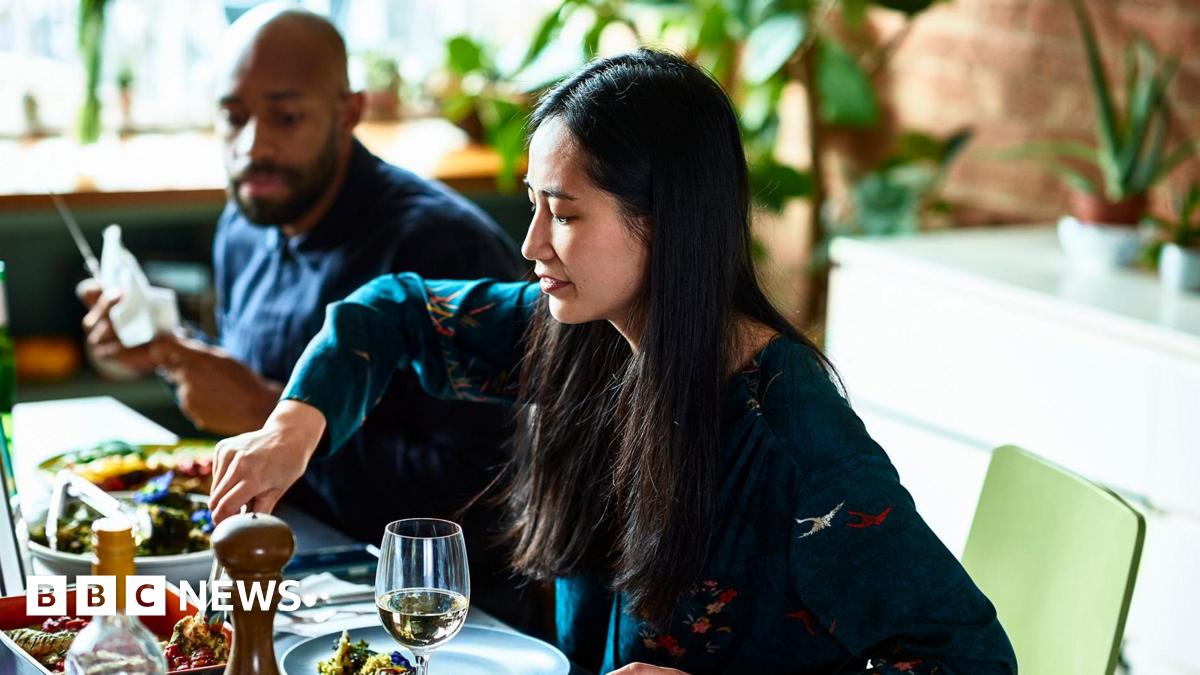 A woman and man sit side-by-side with plates of food in front of them. The woman is serving herself from a dish in front of her.
