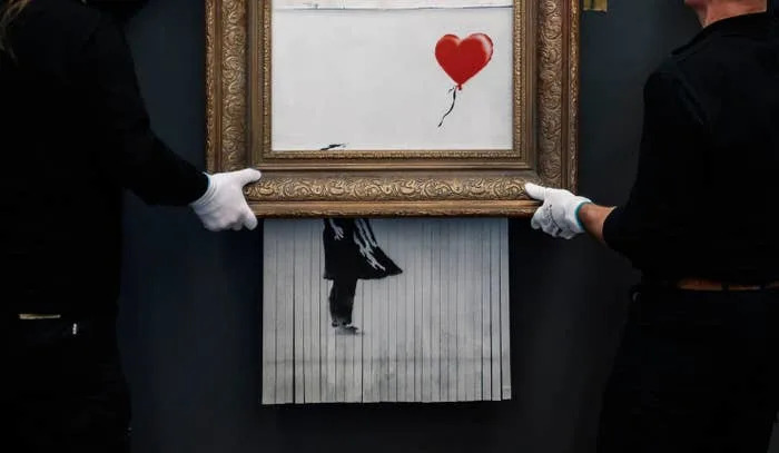 Two people hold a picture frame with shredded artwork below a girl and balloon design