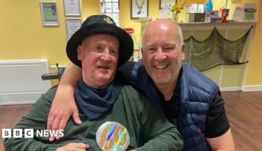 Anthony has grey hair and beard in a blue gilet with a black t-shirt underneath. He has his arm round his brother Keith who is in a wheelchair and is wearing a green long-sleeved t-shirt with a navy bandana round his neck and a black hat on.