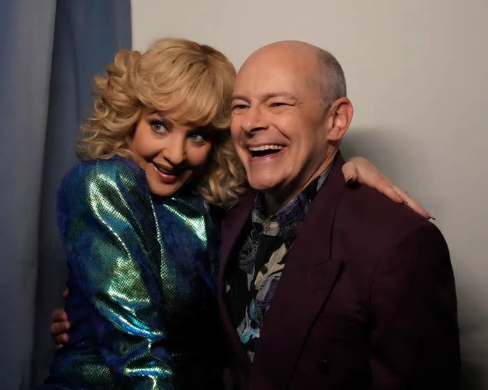 Two people smiling and hugging, one wearing a shimmering jacket and the other in a patterned shirt and dark blazer