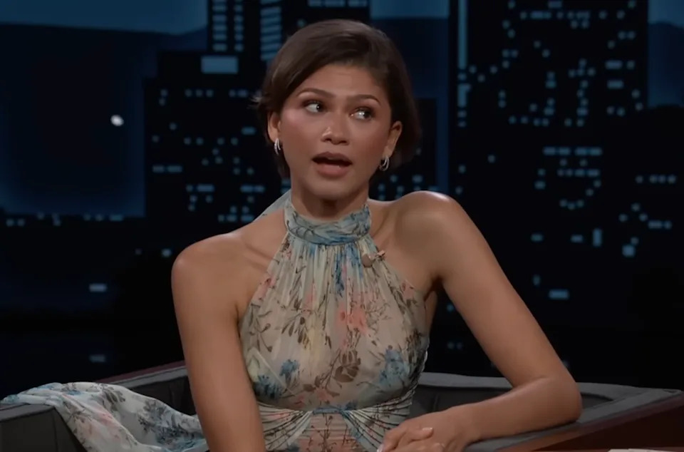 Person in floral halter dress appears to be speaking during a TV interview, seated against a backdrop of a city silhouette
