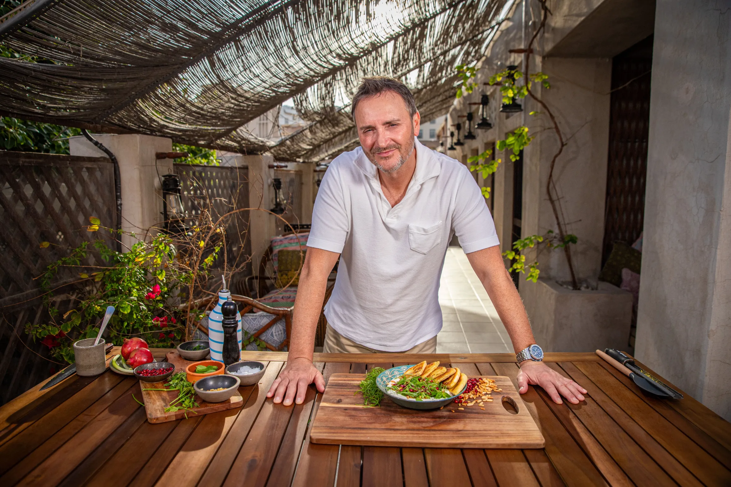 Chef Jason Atherton stands behind a prepared dish, ingredients laid out on a wooden table.