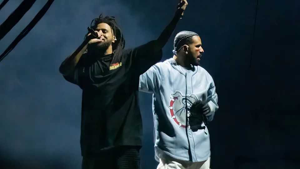 Two performers on stage, J. Cole with a microphone and dreadlocks, the Drake in a light blue jersey, under dramatic lighting.