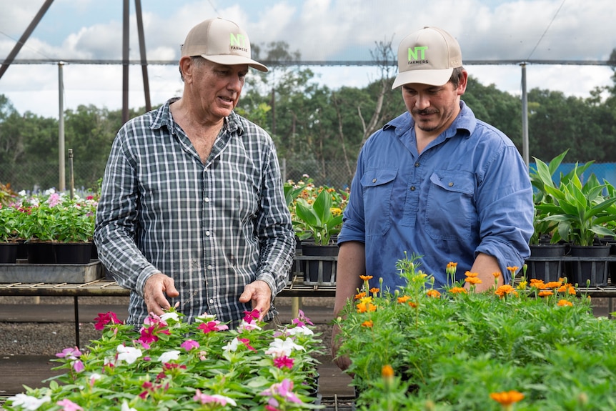 Two men in light coloured caps, one in a checked shirt, the other in a blue shirt stand in front of a bed of colourful flowers