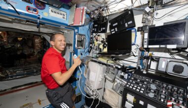 NASA astronaut Chris Williams is seated inside the Destiny laboratory module aboard the International Space Station. He is wearing a red shirt and black shorts with three white stripes underneath the right pocket. Chris is holding a device with a cord in his left hand and there are blank computer screens in front of him. He is smiling brightly and looking directly at the camera.