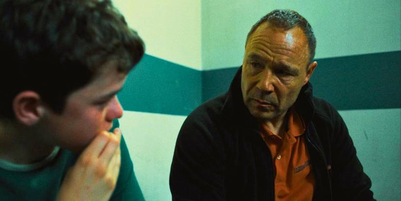 Stephen Graham wears an orange shirt with a black sweater over it and talks to a young boy inside a police station