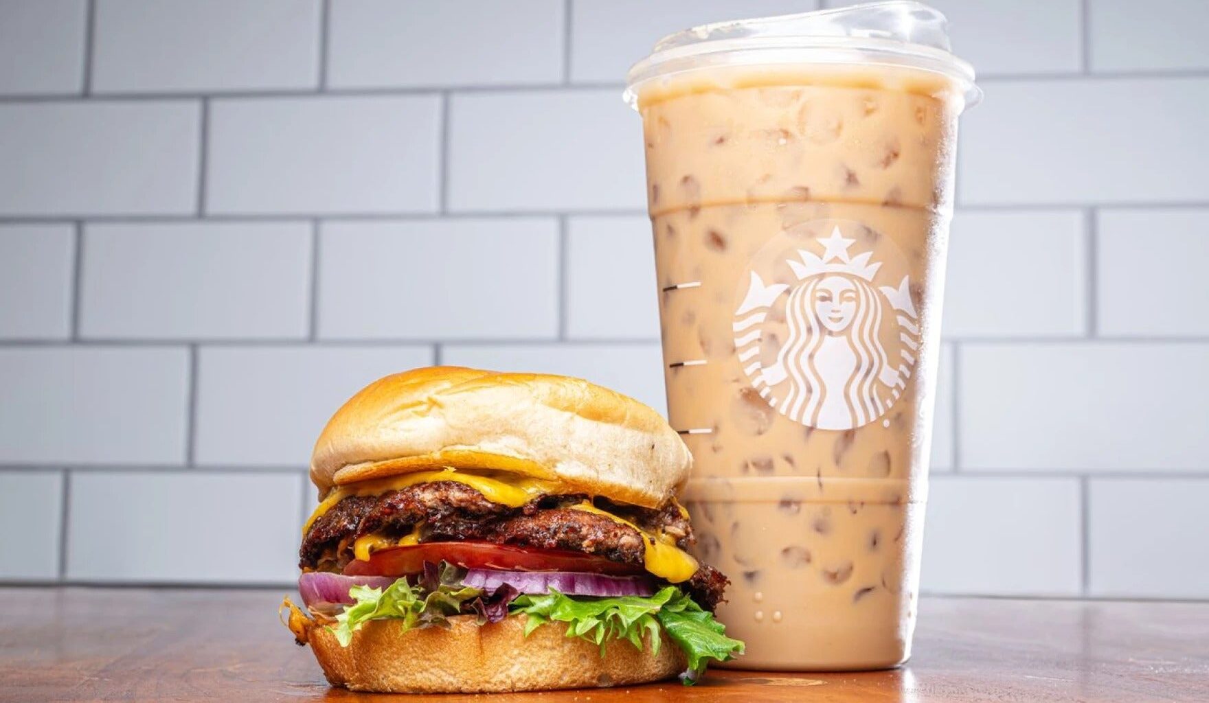 A cheeseburger with lettuce, tomato, onions, and pickles sits next to a large iced coffee in a clear Starbucks cup on a wooden surface.