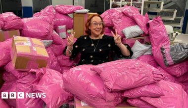 Rachel Grady sits among a large pile of pink parcels. She has a ginger curled bob and holds two peace signs up to either side of her head. She smiles widely and wears pink glasses.