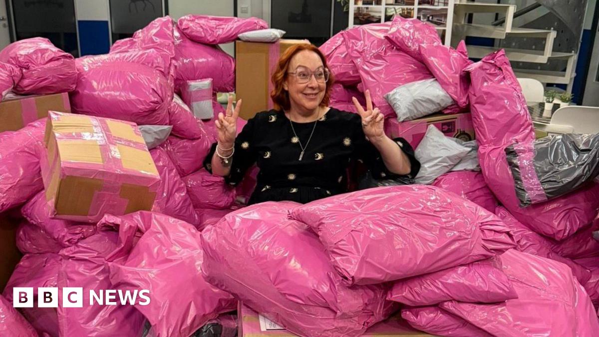 Rachel Grady sits among a large pile of pink parcels. She has a ginger curled bob and holds two peace signs up to either side of her head. She smiles widely and wears pink glasses.