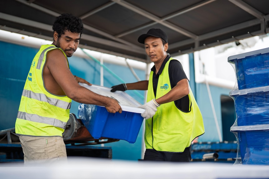 Two men in yellow vests hold either side of a large blue tub
