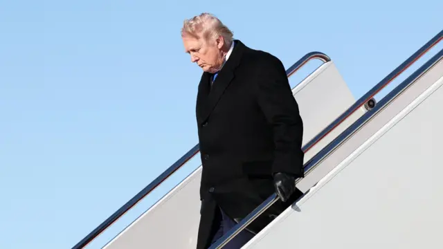 President Donald Trump disembarking a plane dressed in a black coat and black gloves, looking down and holding onto the railing.