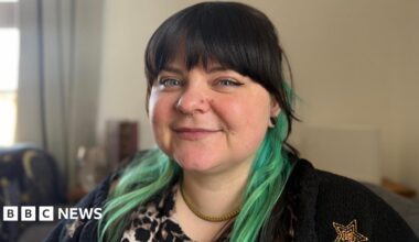 A close-up shot of a woman with a black fringe and teal coloured hair. The lady has several nose piercings and is wearing black.