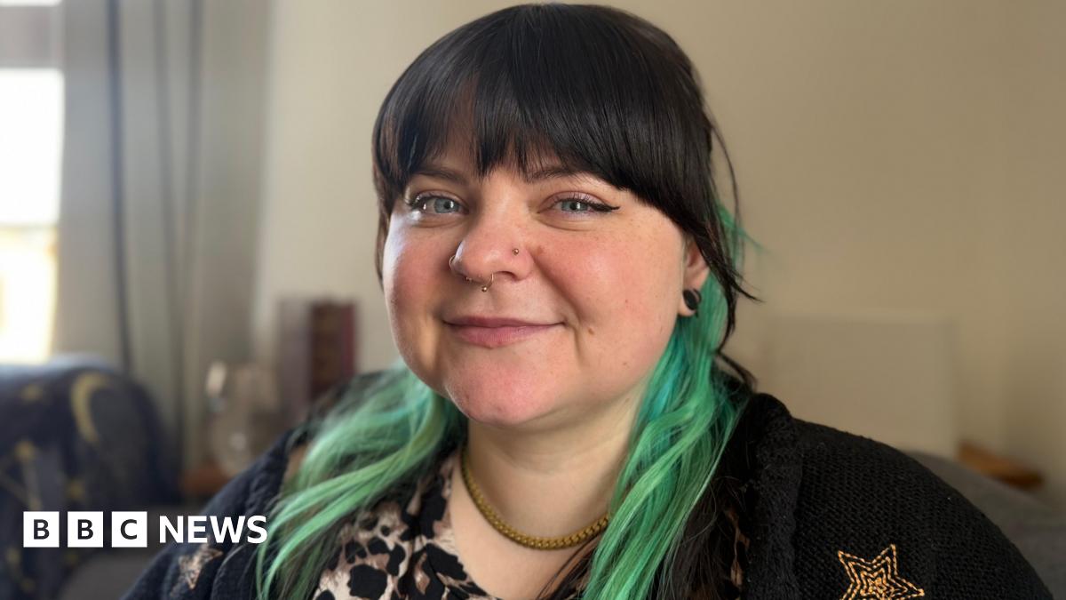 A close-up shot of a woman with a black fringe and teal coloured hair. The lady has several nose piercings and is wearing black.