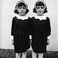 Two young girls, likely twins, stand side by side in matching black dresses and white collars. They wear headbands, with straight-faced expressions. Black and white photo