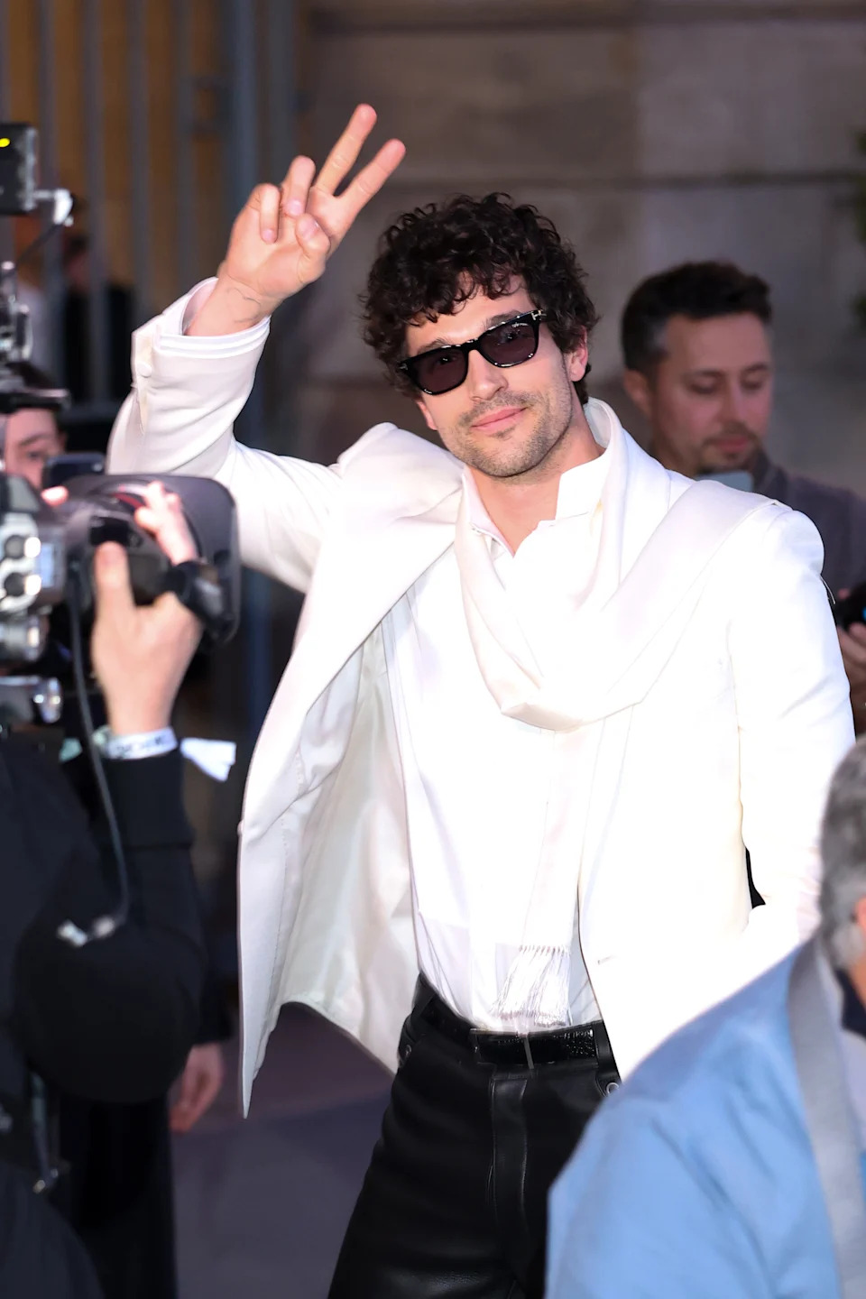 Celebrity on red carpet, wearing a stylish white jacket and sunglasses, making a peace sign with fingers while smiling at cameras