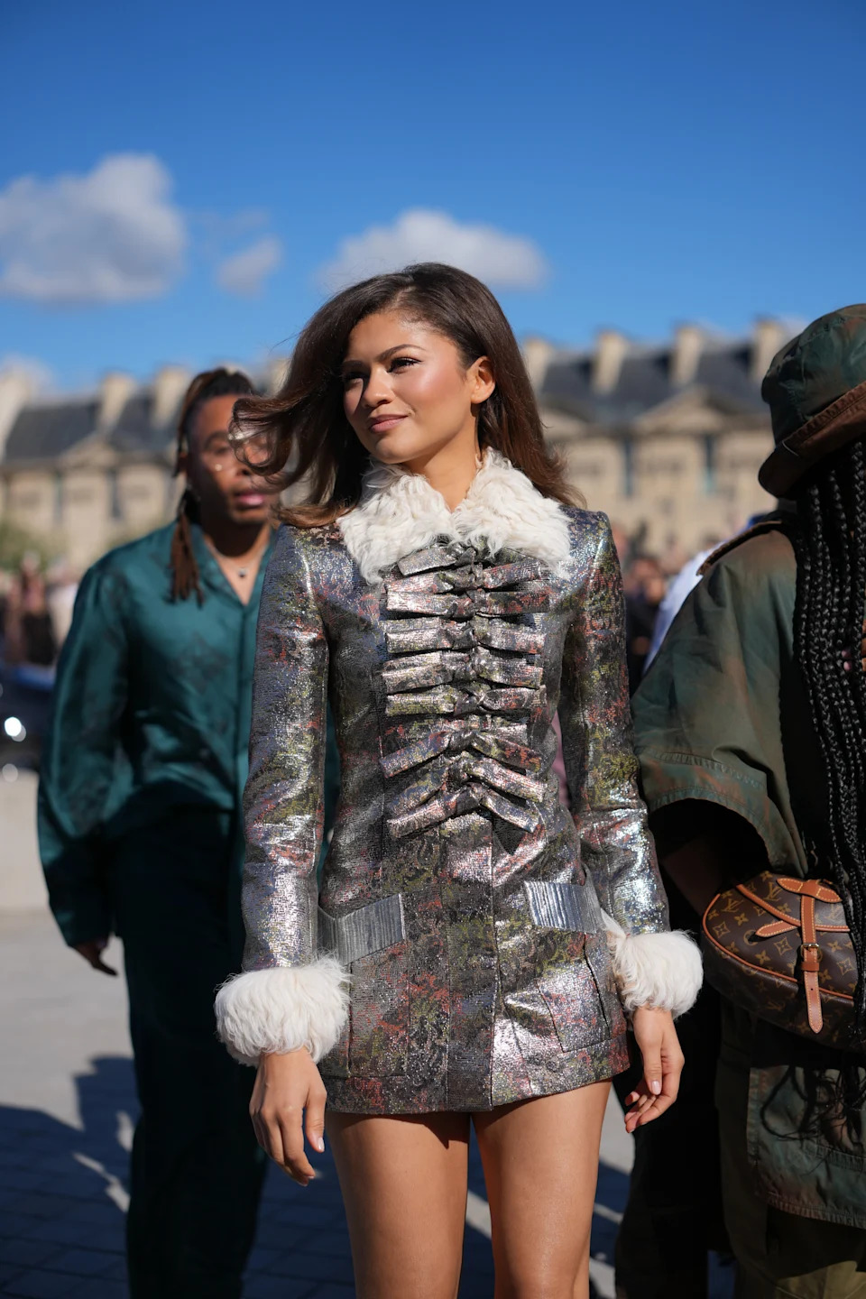 A woman in a metallic patterned jacket and skirt with fur cuffs and collar walks outdoors, followed by other individuals