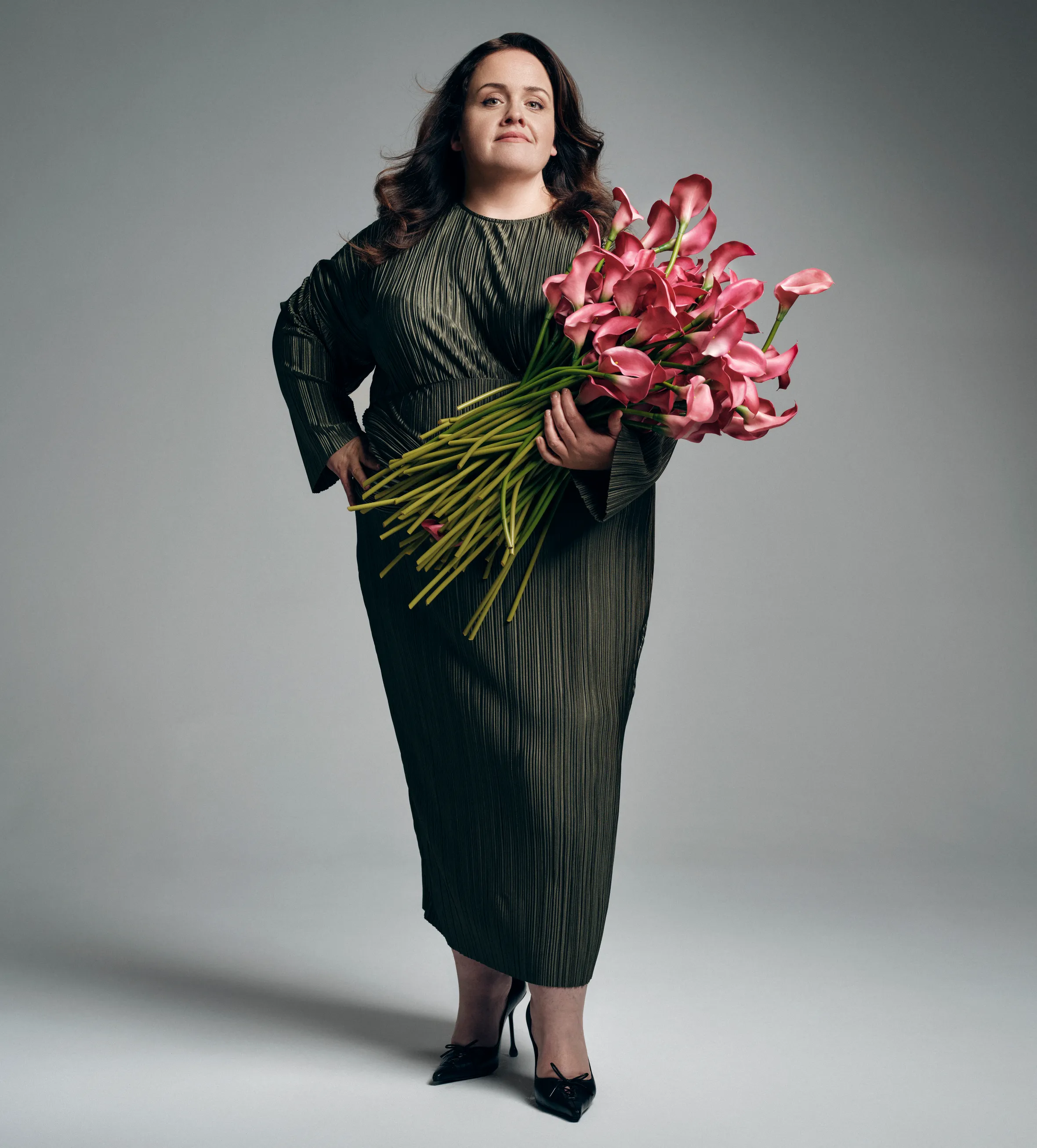 Portrait of Jessica Gunning, wearing a pleated dark dress, holding a large bouquet of pink calla lilies.