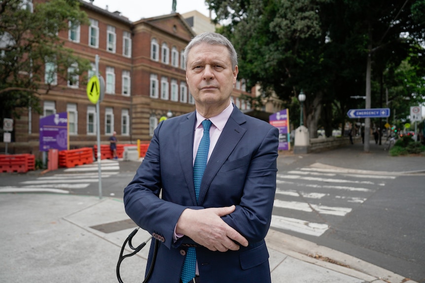 A man in a suit, holds a stethoscope while standing with his arms crossed outside a hospital, with a serious expression.