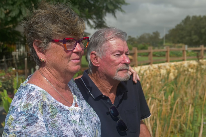 Older woman and man stand with looking into distance with reeds and a path in background.