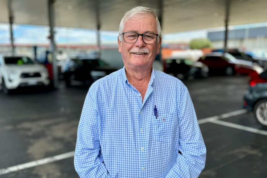 Peter Anderson wear blue and white check shirt. Petrol station behind him.