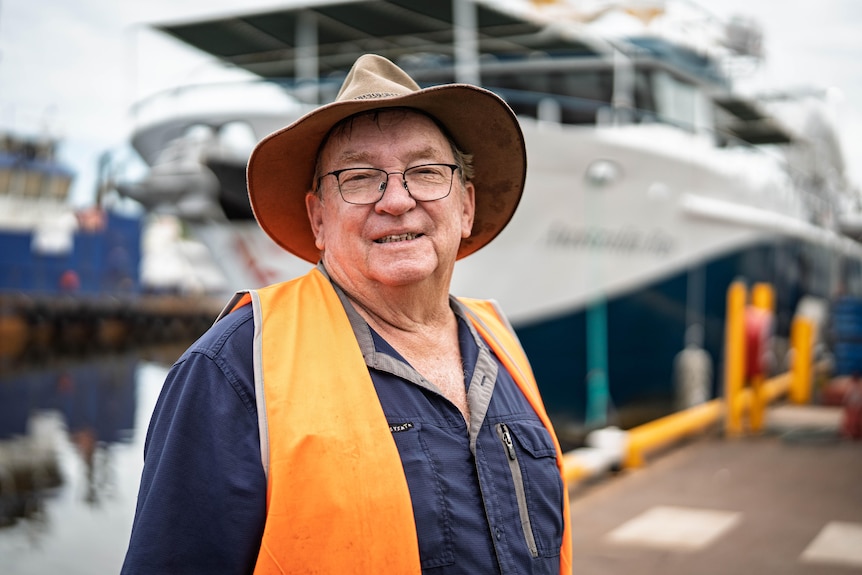 A man in a brown hat with and orange vest over the top of a navy work shirt smiles at the camera