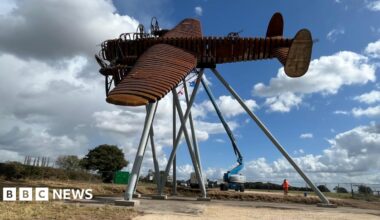 A life-size steel brown sculpture of a Lancaster bomber in a flying motion and tilted to the side. It is held up high by several posts and construction vehicles and a person in orange hi-vis can be seen next to it.
