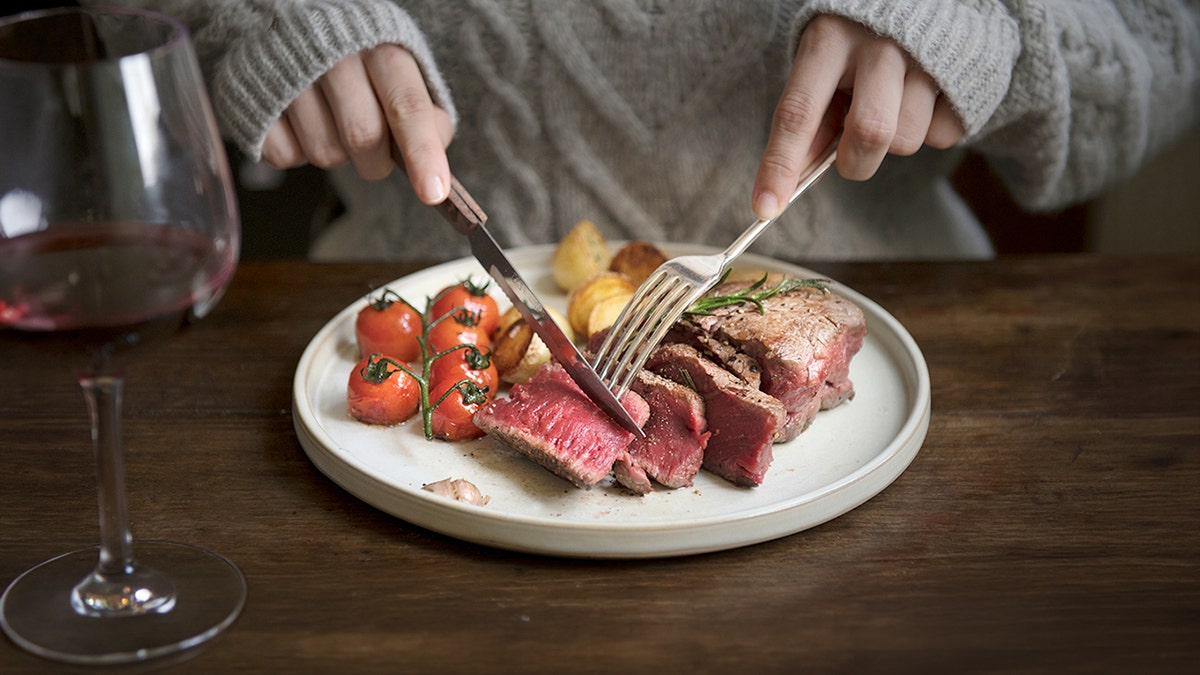 A knife and fork are used to cut into red meat on a plate.
