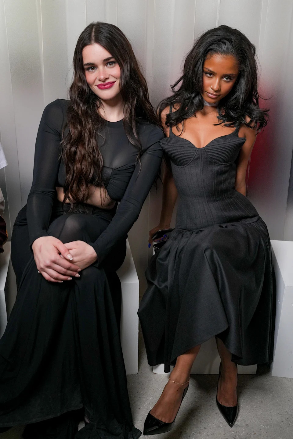 Two women in elegant black dresses, seated and smiling at a formal event. The dresses feature stylish silhouettes