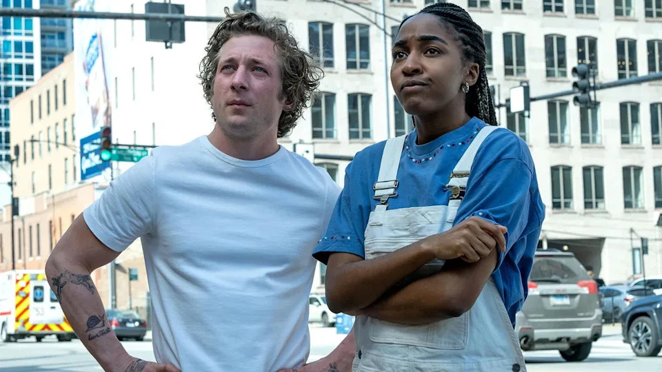 Jeremy Allen White and Ayo Edebiri stand on a city street, looking up. One wears a white shirt, the other a blue shirt with white overalls. Buildings are in the background.