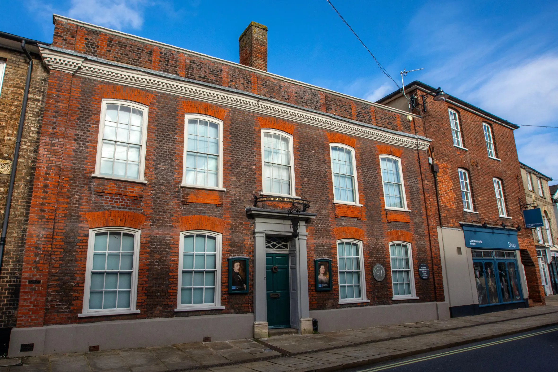 Exterior of Gainsborough's House in Sudbury, Suffolk, a red brick building with white windows, a dark green front door, and signs referencing Thomas Gainsborough.