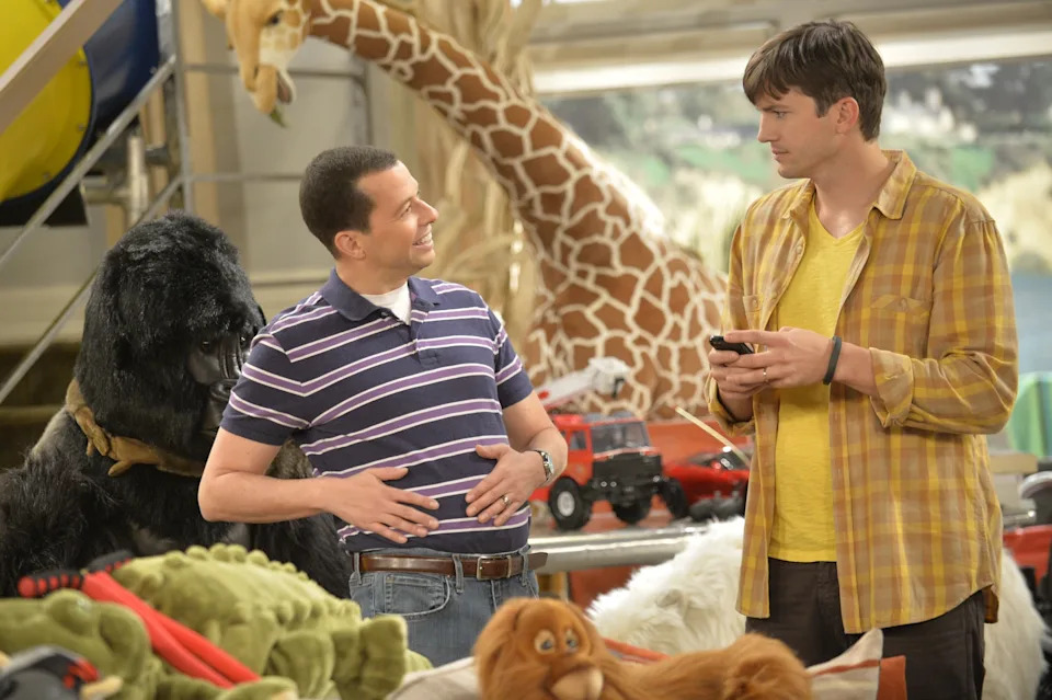 Two men in a room filled with stuffed animals and toys, conversing. One wears a striped polo; the other a plaid shirt, holding a phone