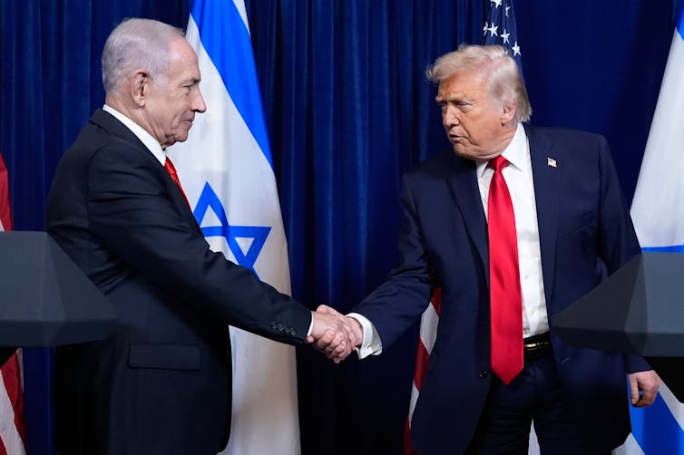 Two old white men in suits shake hands in front of the Israeli and American flags