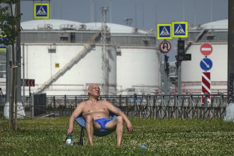 A man in a sun lounger and a Speedo sunbathes in front of a bank of oil storage tanks.