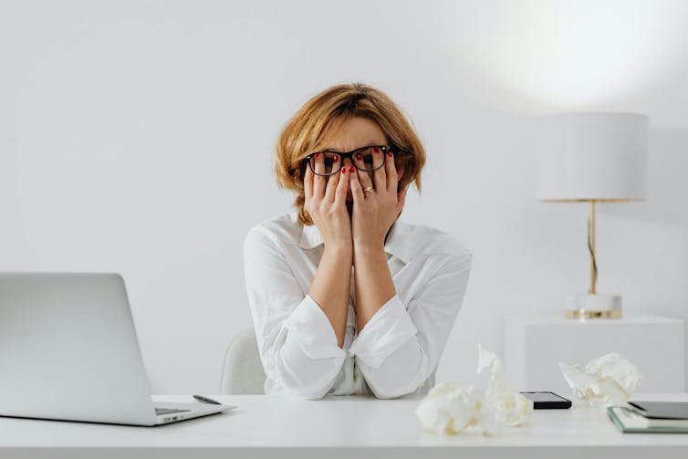 A woman in a white blouse at a desk with a laptop and crumpled papers, burying her face in her hands
