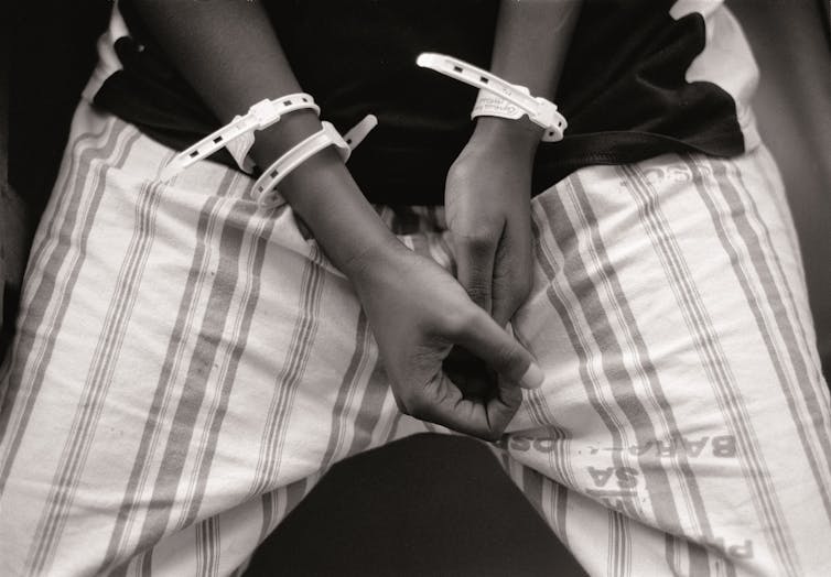 A black and white photo of a person holding their hands together, medical tags on their arms.