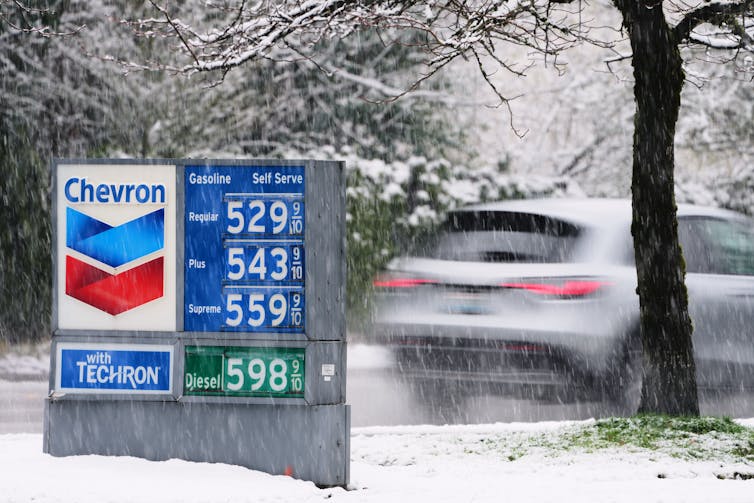 a sign listing high gas prices can be seen as a car drives away in the snow