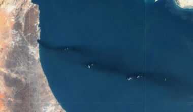 Most of the image is the deep blue of the Persian Gulf, to the left a strip of brown and orange land curves downwards, a third of the way down a dark plume is visible stretchin all the way across the sea to the right of the image.