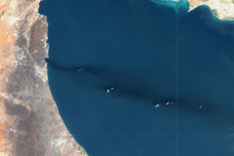 Most of the image is the deep blue of the Persian Gulf, to the left a strip of brown and orange land curves downwards, a third of the way down a dark plume is visible stretchin all the way across the sea to the right of the image.