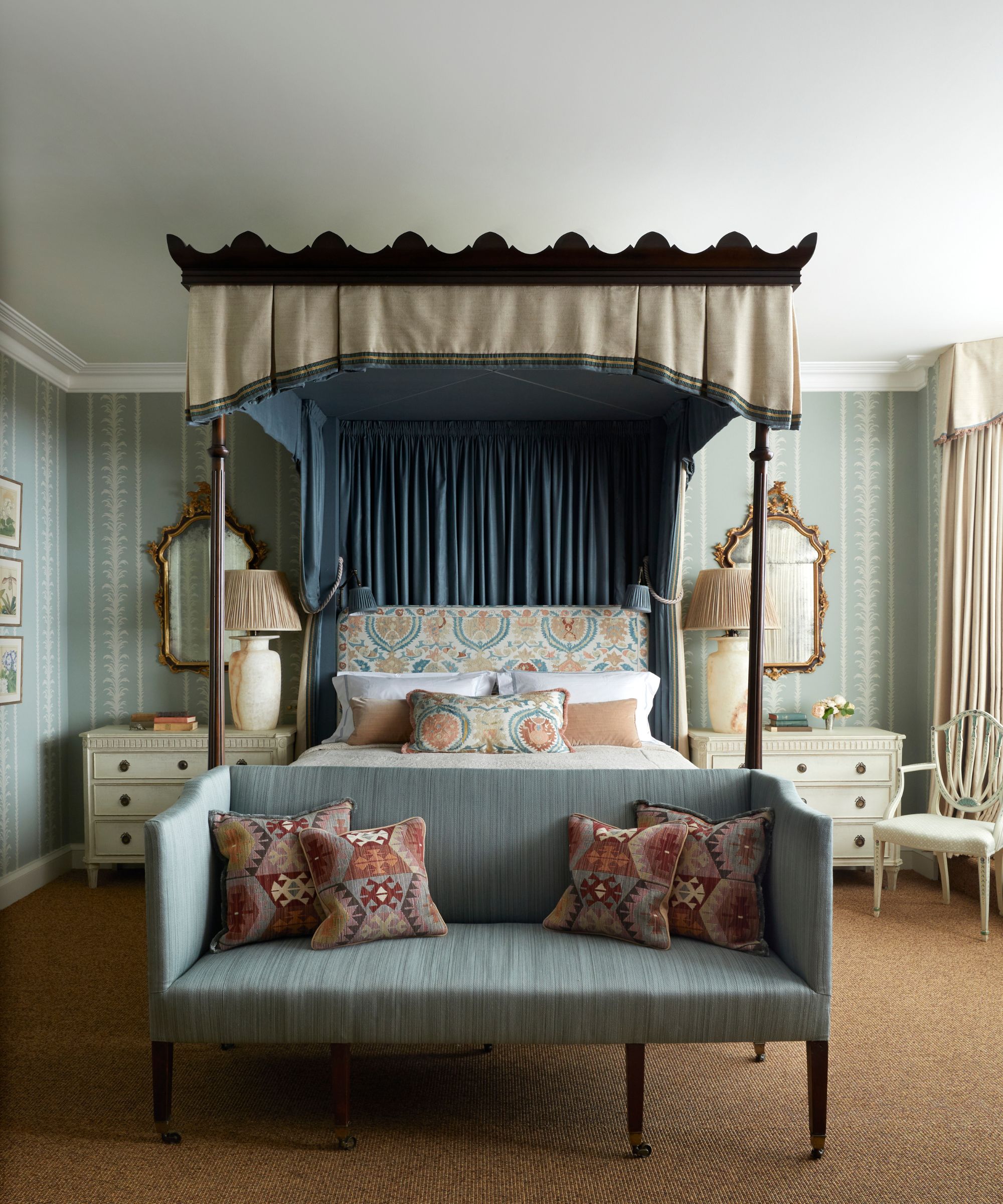 Traditional bedroom with wallpapered walls, jute carpet, four-poster bed with floral headboard, blue bench at the end, and rococo-style white nightstands and gold mirrors above