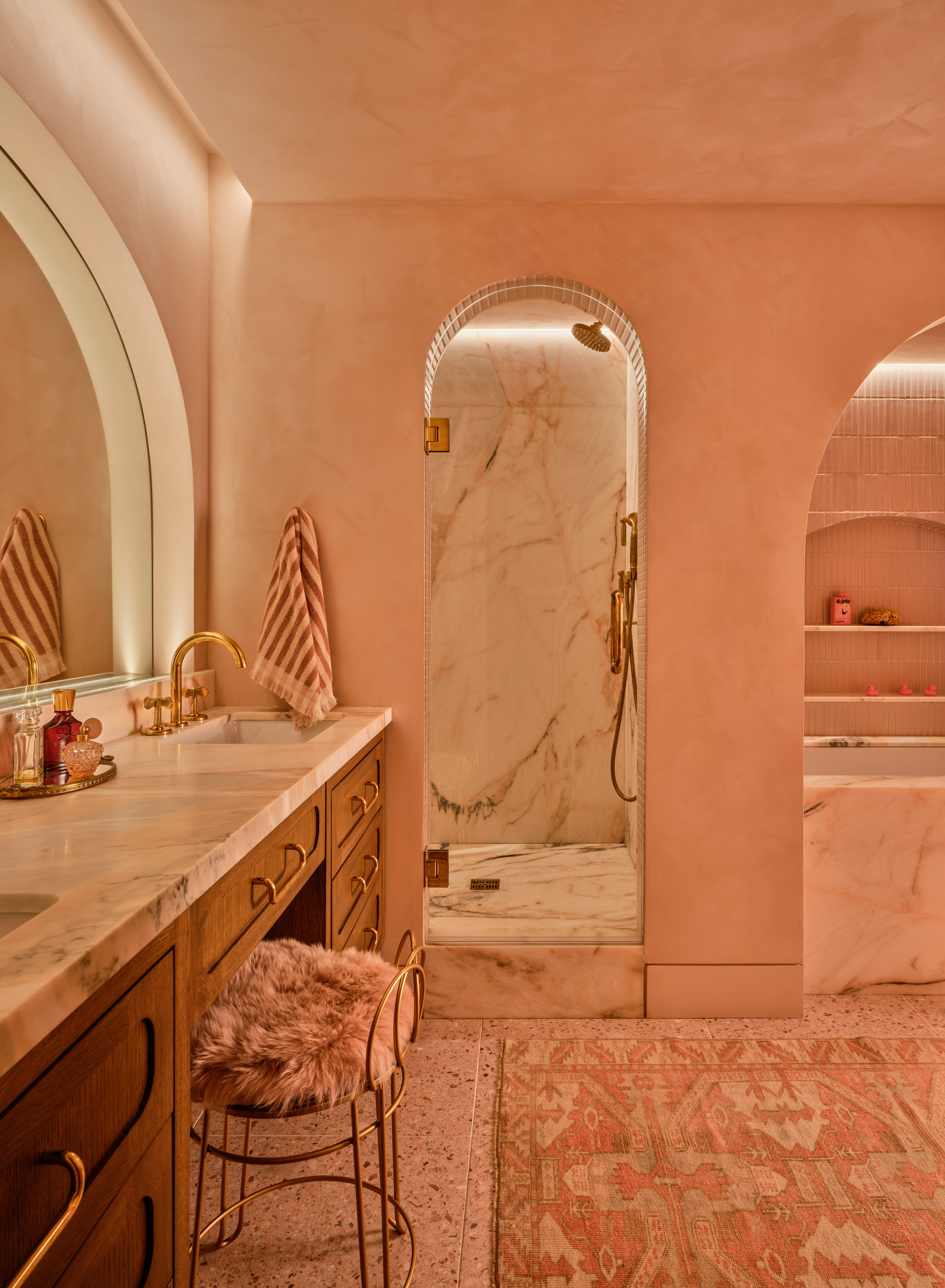 Image of a luxurious bathroom in a pink/peach color drench with a double sink, walk-in shower, pink rug, and a fuzzy pink stool at the vanity area.