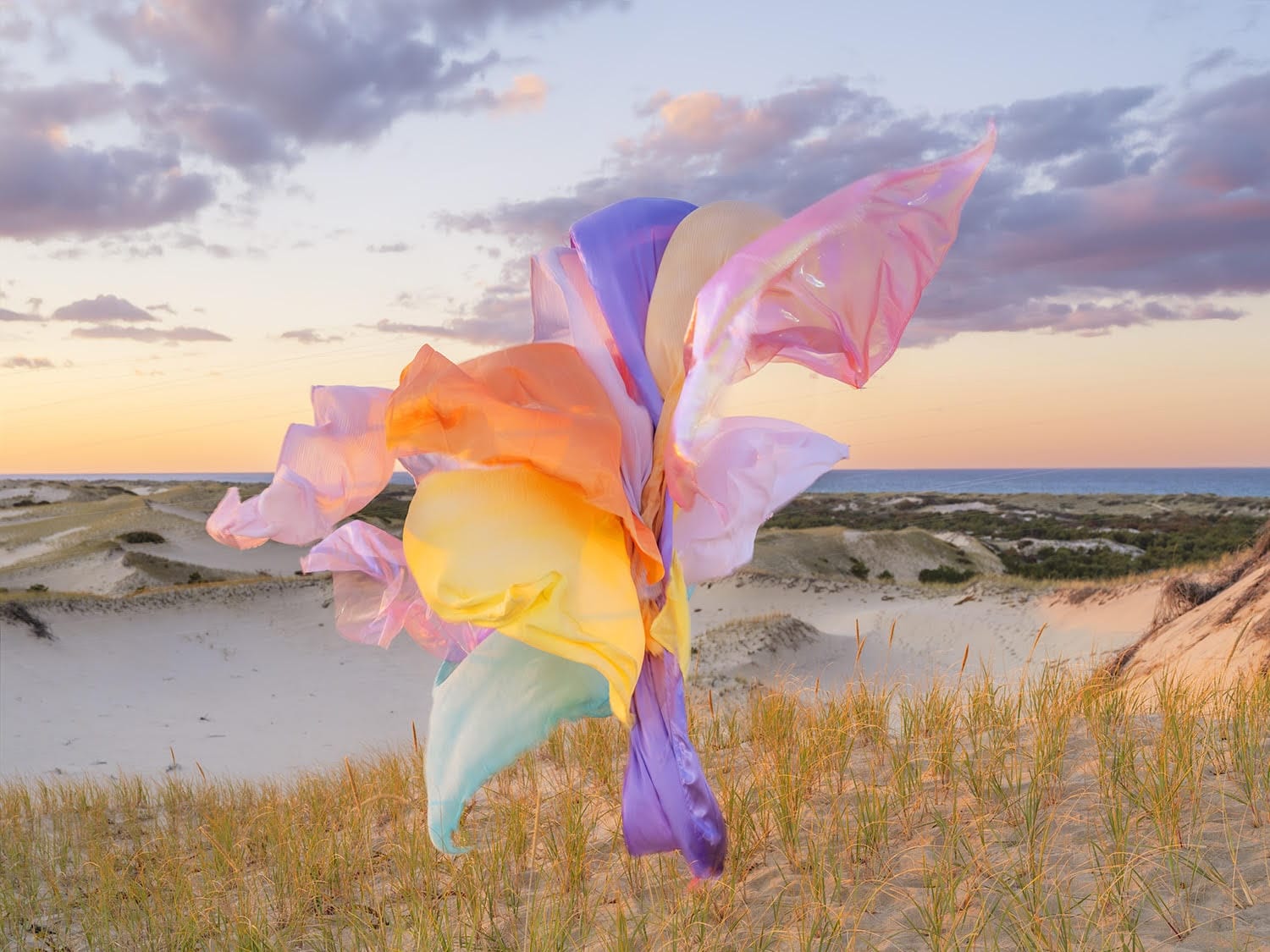 A photograph by Thomas Jackson of numerous pieces of colorful fabric that are seemingly suspended in the air above a meadow near a lake