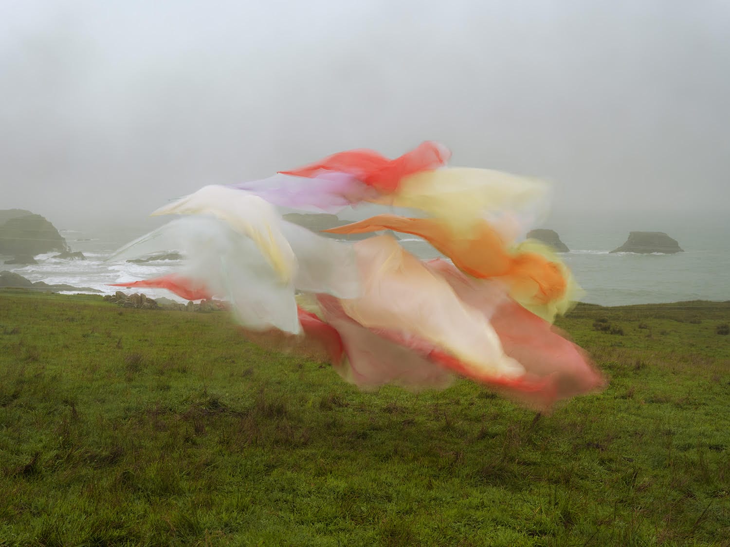 A photograph by Thomas Jackson of numerous pieces of colorful fabric that are seemingly suspended in the air above a meadow