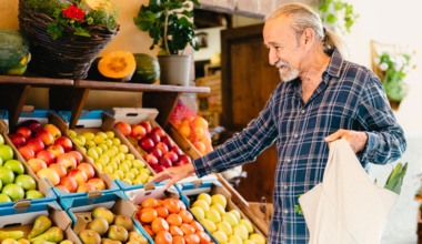 Elderly man in a plaid shirt smiling while selecting fruits from a market stand during National Nutrition Month. Vibrant apples, pears, and tomatoes are displayed in boxes nearby.