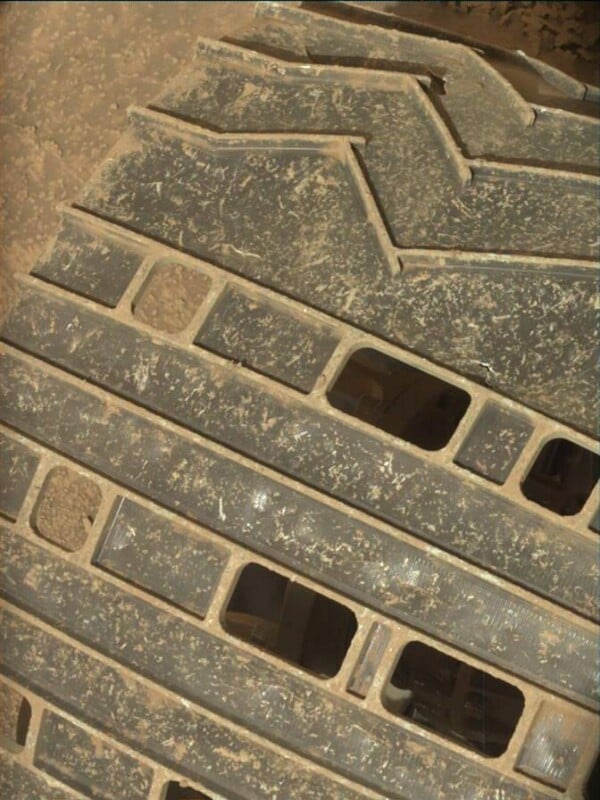 Close-up view of a dusty rover wheel on Mars, showing metal treads with rectangular cutouts and a zigzag pattern. The surface is coated with Martian sand.