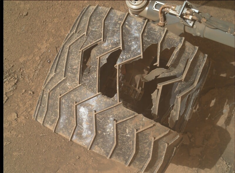 A close-up view of a damaged rover wheel on a dusty, reddish surface. The metal wheel has large cracks and holes, with bent and broken segments, and part of the rover’s suspension arm is visible in the background.