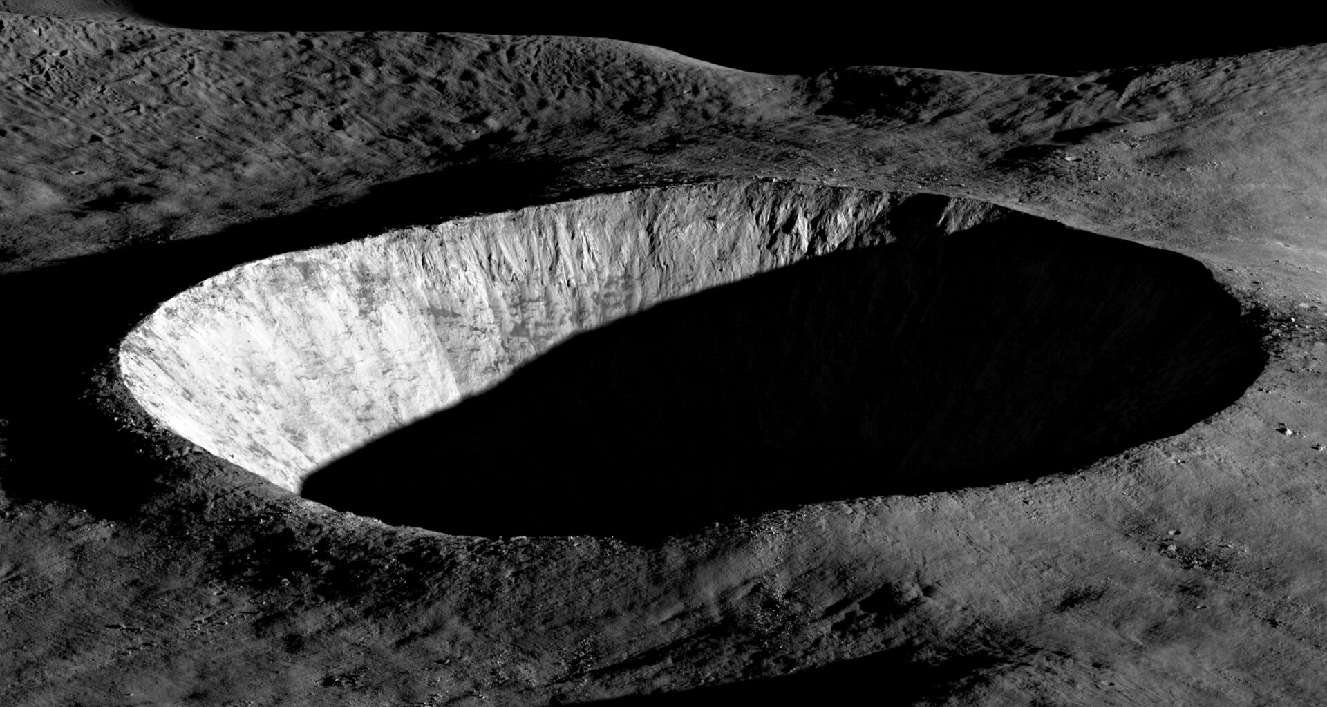 Oblique view of a large, circular lunar crater with steep, rugged walls. Sunlight illuminates one side of the crater rim and inner wall, while the crater floor and much of the interior remain in deep shadow. The surrounding terrain appears gray and heavily textured with smaller craters and rocky ridges. The black sky of space fills the background.