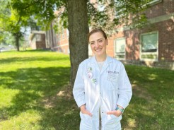 A person wearing a white lab coat stands outside on grass near a brick building and trees, smiling with hands in their pockets. Their name badge reads “Acadia Hospital.” A pin promoting lemon eucalyptus oil as tick repellent is on the lab coat.