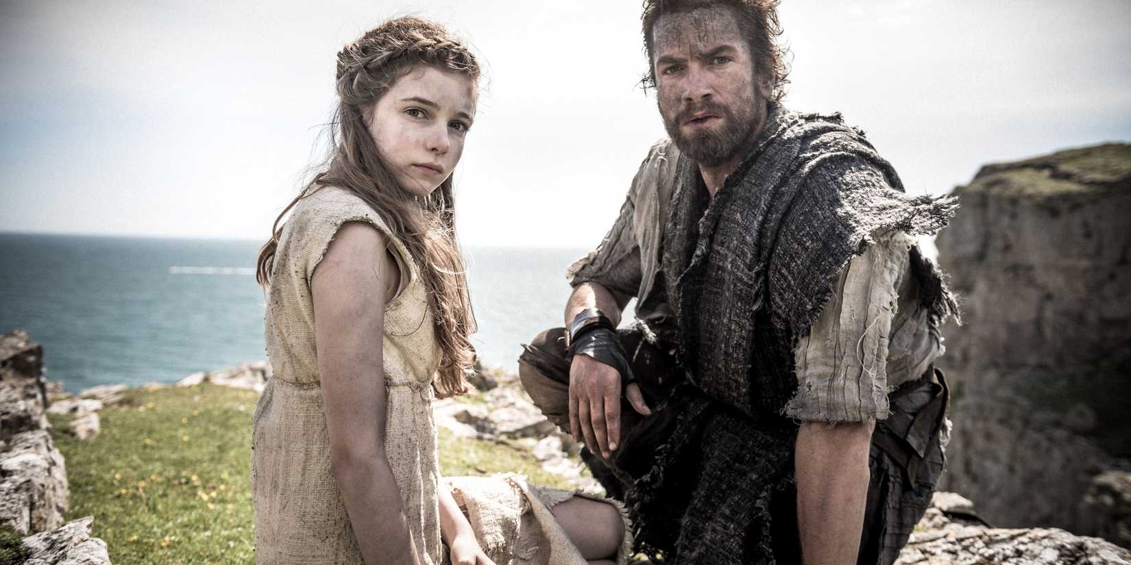 Nikolaj Lie Kaas as Divis, and Eleanor Worthington-Cox as Cait, staring directly at the camera, sitting atop a cliffside with an ocean in the background, in Britannia