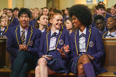 Three students in school uniforms sit together in an assembly and laugh (Xiang, Mabel, Devoyne - Boarders) 